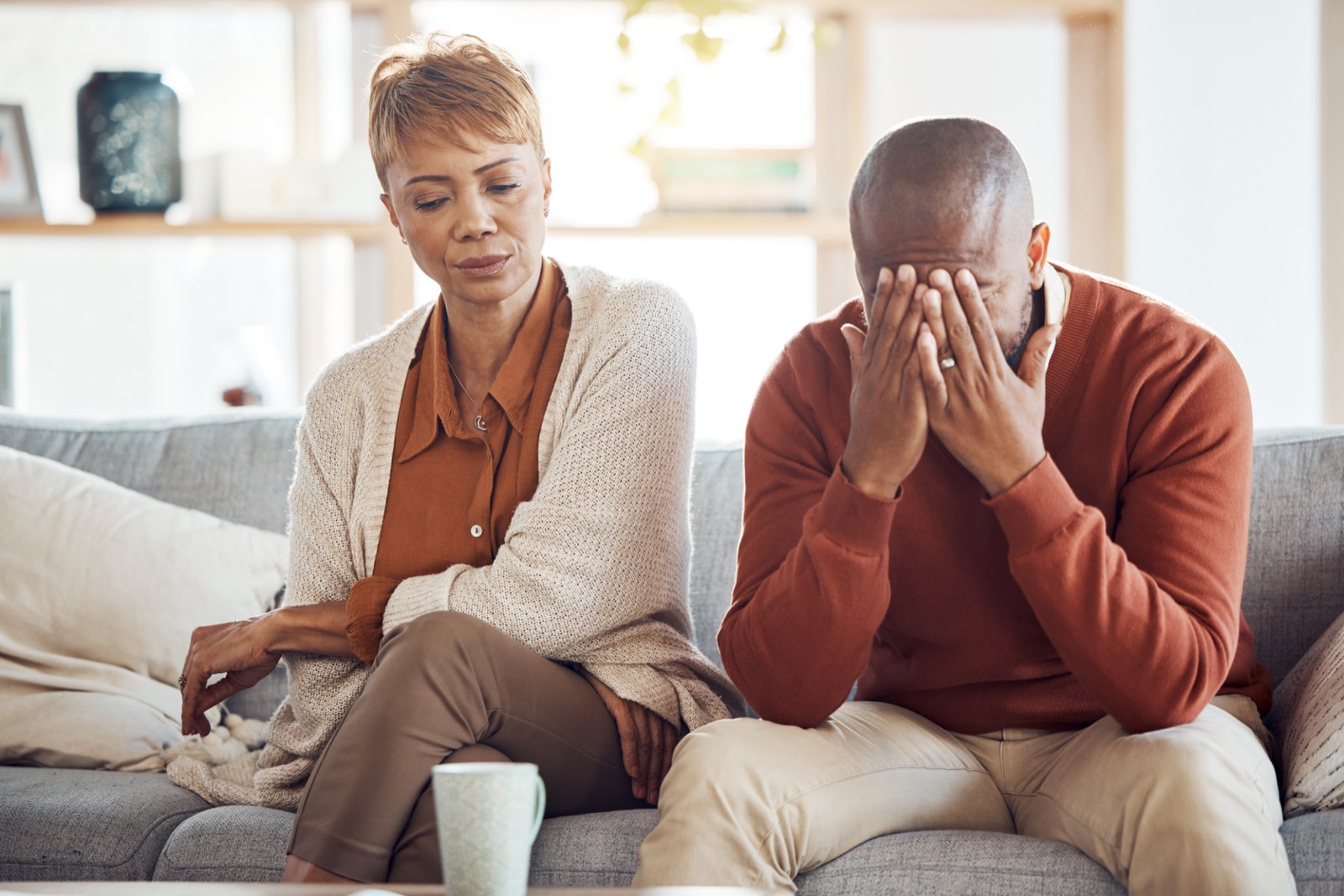 A couple sitting on a sofa, with one person covering their face with their hands.